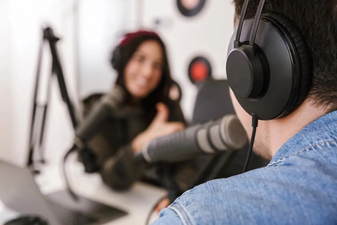 Audio-Aufnahme zwei Personen Eine Person sitzend mit Kopfhöhrer spricht in ein Mikrofon. Gegenüber sitz eine Frau ebenfalls mit Kopfhörern.